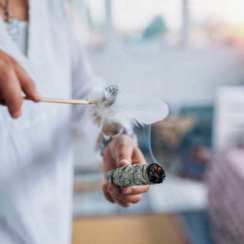 Woman’s hand holding burning sage smudge stick, spreading the smoke around with white feather, blessing and cleansing home from negative energies